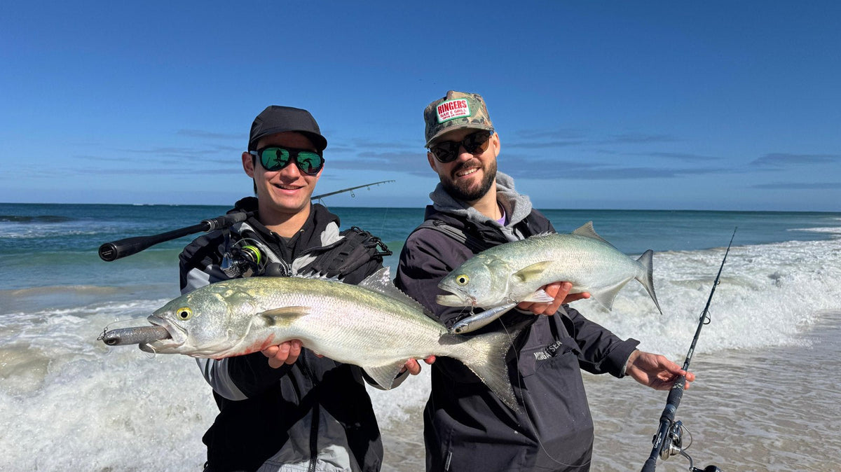 Two anglers standing in the shallows holding tailor caught beach fishing, with surf rods and waves behind them