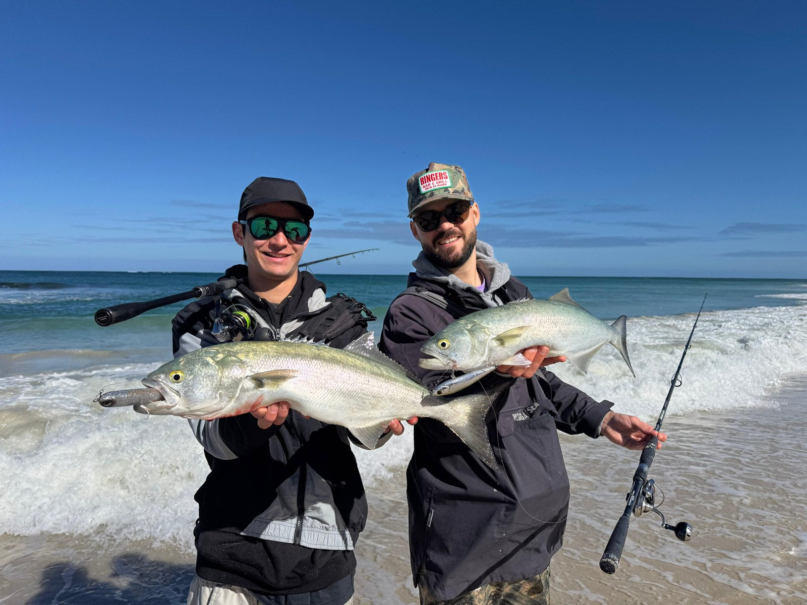 Two anglers standing in the shallows holding tailor caught beach fishing, with surf rods and waves behind them